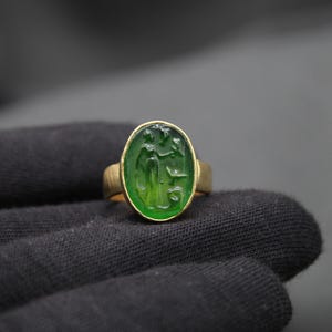 May include: A gold ring with a green oval gemstone featuring a carved design. The ring is set on a black fabric background.