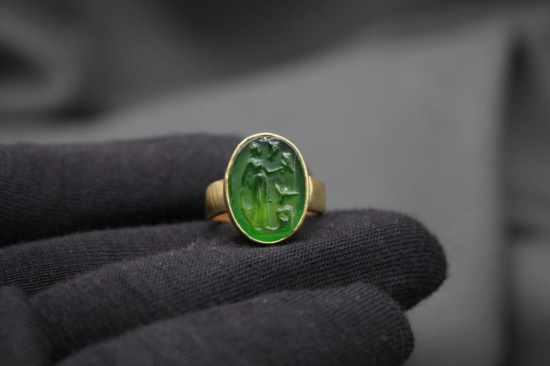 May include: A gold ring with a green oval gemstone featuring a carved design. The ring is set on a black fabric background.