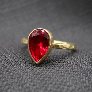 May include: A gold ring with a pear-shaped, vibrant red gemstone. The ring's band has a textured, hammered appearance. The gemstone is set in a gold bezel, creating a striking contrast against the gold band. The ring is displayed on a gray surface.