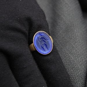 May include: A gold ring with a blue oval-shaped stone featuring a carved design of a woman holding a staff.