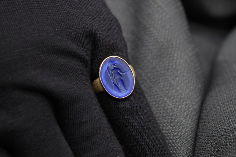 May include: A gold ring with a blue oval-shaped stone featuring a carved design of a woman holding a staff.