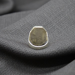 K&ouml;nnte beinhalten: Ein silberner Ring mit einer glatten, ovalen Oberseite. Der Ring liegt auf einem grauen Stoffhintergrund.