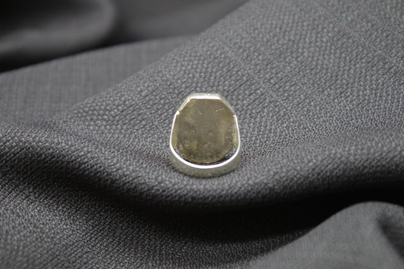 K&ouml;nnte beinhalten: Ein silberner Ring mit einer glatten, ovalen Oberseite. Der Ring liegt auf einem grauen Stoffhintergrund.