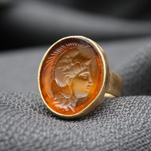 May include: A gold ring with an oval-shaped amber stone featuring a carved image of a woman's head wearing a helmet.