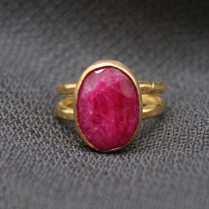 Könnte beinhalten: Ein goldfarbener Ring mit einem großen, oval geformten, tiefrosa Edelstein. Der Ring hat ein Doppelband-Design, wobei der Edelstein in einer goldenen Lünette gefasst ist. Der Ring ist vor einem dunkelgrauen, strukturierten Hintergrund platziert.