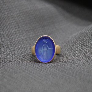 May include: A gold ring with a blue oval shaped stone featuring a carved design of a woman holding a staff.