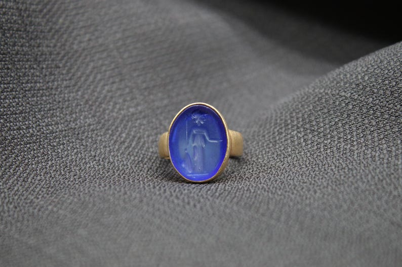 May include: A gold ring with a blue oval shaped stone featuring a carved design of a woman holding a staff.