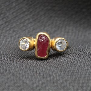 Könnte beinhalten: Ein goldener Ring mit einem roten Edelstein und zwei klaren Edelsteinen.