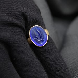 May include: A gold ring with a blue oval-shaped stone featuring a carved design. The ring is on a dark fabric background.
