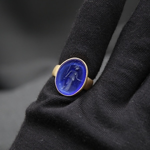 May include: A gold ring with a blue oval-shaped stone featuring an engraved image of a person.