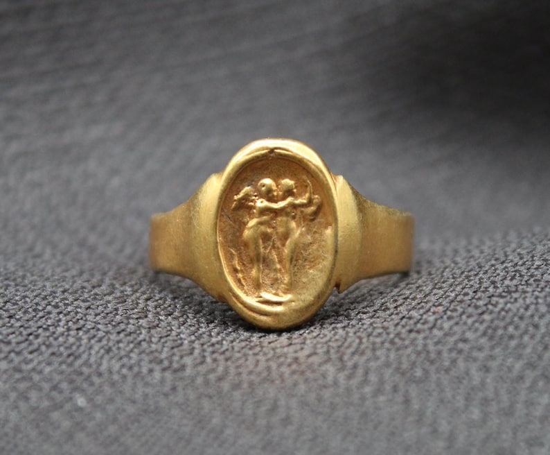 May include: A gold ring with an oval-shaped design featuring a raised image of two figures embracing. The ring has a smooth, polished finish.