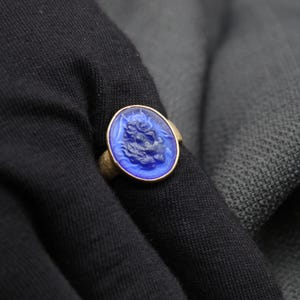May include: A blue glass ring with a detailed design of a face. The ring is set in a gold band.