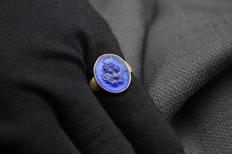 May include: A blue glass ring with a detailed design of a face. The ring is set in a gold band.