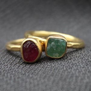 Könnte beinhalten: Zwei goldene Ringe mit einem roten Rubin und einem grünen Smaragd. Die Ringe sind übereinander gestapelt.