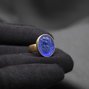 May include: A gold ring with a blue oval-shaped stone featuring a carved design. The stone is set in a gold band.