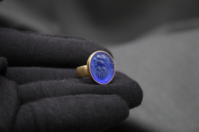 May include: A gold ring with a blue oval-shaped stone featuring a carved design. The stone is set in a gold band.