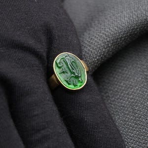 May include: A gold ring with a green stone featuring a carved design. The stone is round and has a raised edge.