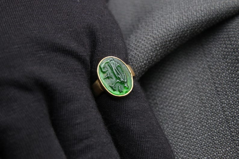 May include: A gold ring with a green stone featuring a carved design. The stone is round and has a raised edge.