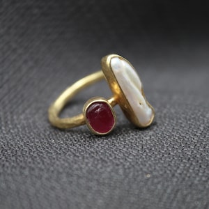 Puede incluir: Un anillo de oro con una perla blanca y una piedra roja. El anillo est&aacute; sobre un fondo de tela gris.
