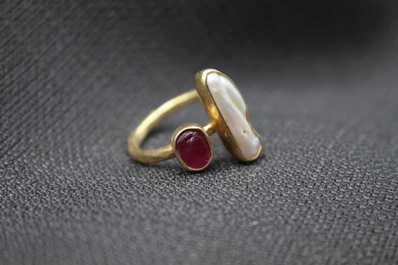 Puede incluir: Un anillo de oro con una perla blanca y una piedra roja. El anillo est&aacute; sobre un fondo de tela gris.