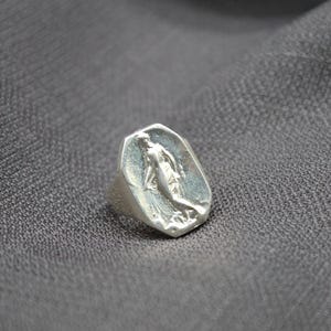 K&ouml;nnte beinhalten: Ein silberner Ring mit einer achteckigen Fl&auml;che, die ein erhabenes Design einer stehenden Frau mit ausgestreckten Armen zeigt.