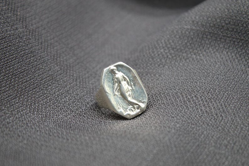 K&ouml;nnte beinhalten: Ein silberner Ring mit einer achteckigen Fl&auml;che, die ein erhabenes Design einer stehenden Frau mit ausgestreckten Armen zeigt.