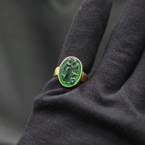 May include: A gold ring with a green oval-shaped stone featuring a carved design. The ring is on a black fabric background.