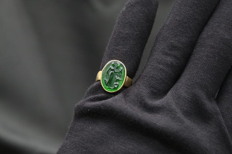 May include: A gold ring with a green oval-shaped stone featuring a carved design. The ring is on a black fabric background.