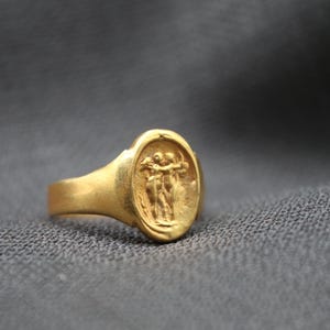 May include: A gold ring with an oval-shaped setting featuring a raised design of two figures embracing.