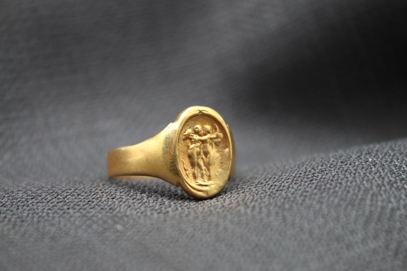 May include: A gold ring with an oval-shaped setting featuring a raised design of two figures embracing.