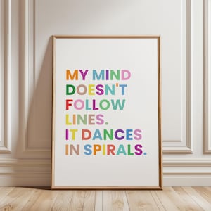 Könnte beinhalten: Ein gerahmter Kunstdruck mit dem Text "MY MIND DOESN'T FOLLOW LINES. IT DANCES IN SPIRALS." Der Text ist in verschiedenen leuchtenden Farben gehalten, darunter Orange, Grün, Rot, Gelb, Lila und Blau. Der Rahmen ist hellbraun.