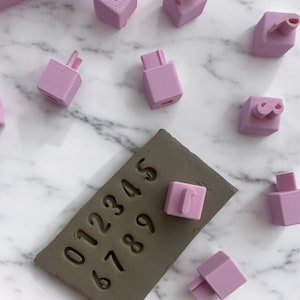 Op de afbeelding: Een set roze siliconen stempels met de cijfers 0-9, een roze siliconen bakje met 12 vierkante vormen en een bruine klei plaat met de cijfers 0-9 erop gedrukt.