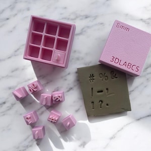 Op de afbeelding: Een set roze alfabetstempels in een roze opbergdoos. De stempels zijn 8 mm groot en bevatten letters, cijfers en symbolen. De stempels zijn gemaakt door 3DLABCS.