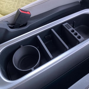 May include: A black car console organiser with a cup holder, dividers, and a phone holder. The organiser is designed to fit inside a car's centre console and keep items organised. The interior is black with a silver trim.