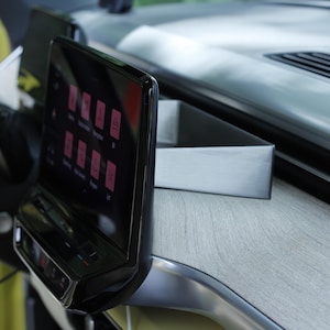 May include: A car interior detail featuring a black touchscreen with pink icons. The dashboard has a light wood effect and silver trim. A steering wheel is visible on the left side of the image.