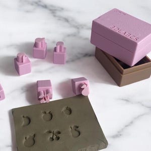 May include: A set of pink plastic stamps in a pink and brown box. The stamps feature different fruit designs, including a pear, apple, cherry, and a crescent moon. The stamps are used to create impressions in a brown clay surface.