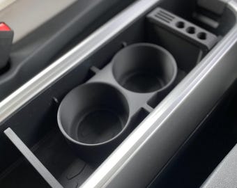 Op de afbeelding: Een zwarte bekerhouder voor de auto console met twee ronde openingen. De bekerhouder is geplaatst in een groter compartiment met een zilveren rand. Het interieur van de auto is zichtbaar.
