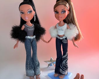 May include: Two fashion dolls with high ponytails. The left doll wears a grey tank top with the word Sugar, a black faux fur shrug, and rhinestone-embellished jeans. The right doll wears a white tank top with the word Girly, a white faux fur shrug, and rhinestone-patterned dark denim jeans.