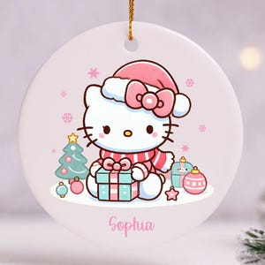 May include: A round Christmas ornament featuring a cartoon cat wearing a Santa hat and scarf, holding a gift. The ornament has a pink and blue color scheme with snowflakes and a Christmas tree. The name "Sophia" is written on the ornament.