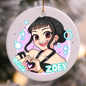 May include: Round ceramic ornament featuring a cartoon illustration of a smiling person with dark hair and red eyes. The person is making a heart shape with their hands. The names "Olivia" and "Zoey" are written on the ornament.