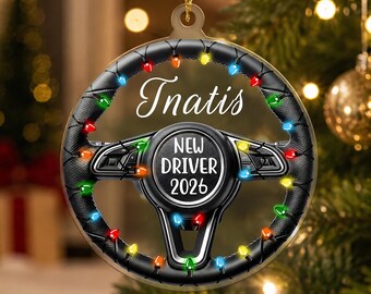 Pode incluir: Um ornamento circular em forma de volante preto com aderência texturizada, decorado com luzes de Natal coloridas. O centro apresenta o texto "NEW DRIVER 2026" e o nome "Inatis" está impresso em letra branca acima do volante.