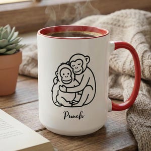 May include: White ceramic mug with a red interior and handle. The mug features a black line drawing of two hugging monkeys and the word "Punch" in a script font. The mug is filled with coffee.