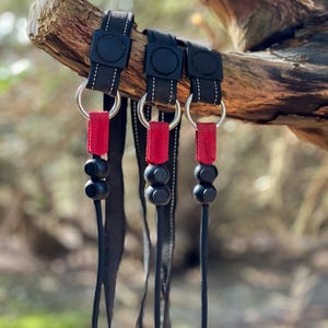 Op de afbeelding: Drie zwarte lanyards met rode en zwarte accenten. Elke lanyard heeft een zwarte ring met een zilveren metalen ring eraan bevestigd. De lanyards hangen aan een tak.