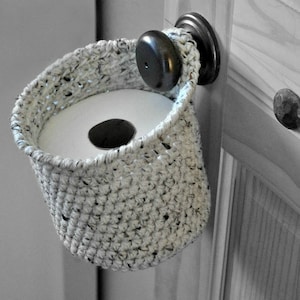 May include: A crocheted toilet paper holder hangs on a door knob. The holder is made of white yarn with black flecks and is shaped like a basket. A roll of white toilet paper is inside the holder.
