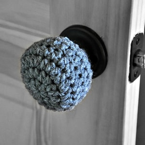 May include: A gray crocheted door knob cover. The cover is made of a soft, textured yarn and is designed to fit over a standard door knob.