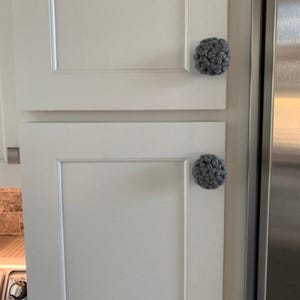 May include: Two white cabinet doors with gray crocheted knobs. The cabinet doors are closed and the knobs are centered on the doors.
