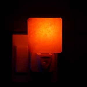 May include: A square, orange night light is plugged into a white wall socket. The night light emits a warm, glowing light. The background is dark, emphasising the illuminated night light. The night light is a decorative home accessory.