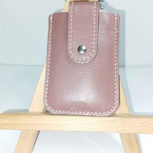 May include: A dusty rose leather card holder featuring a vertical orientation, light-colored contrast stitching along the edges, and a central snap button closure. The rectangular accessory has a small semi-circular cutout at the bottom edge.
