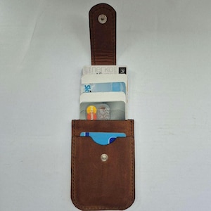 Puede incluir: Un tarjetero de cuero marrón con cierre de botón a presión, que contiene varias tarjetas de crédito. El tarjetero tiene un diseño vertical con una solapa que asegura las tarjetas. La palabra "легко" es visible en una de las tarjetas.