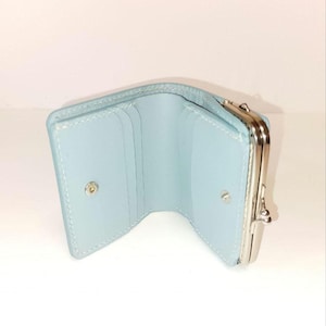 May include: A light blue wallet, open to reveal card slots and a snap closure. It has a silver clasp and is made from a smooth material. The design is minimalist.